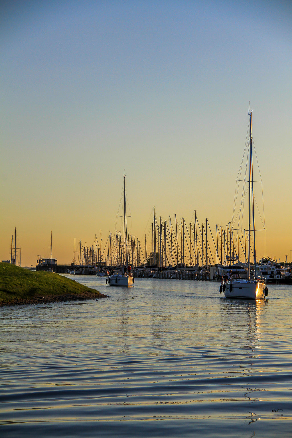 A marina channel at golden hour with sailboats, tall masts and calm rippled water.