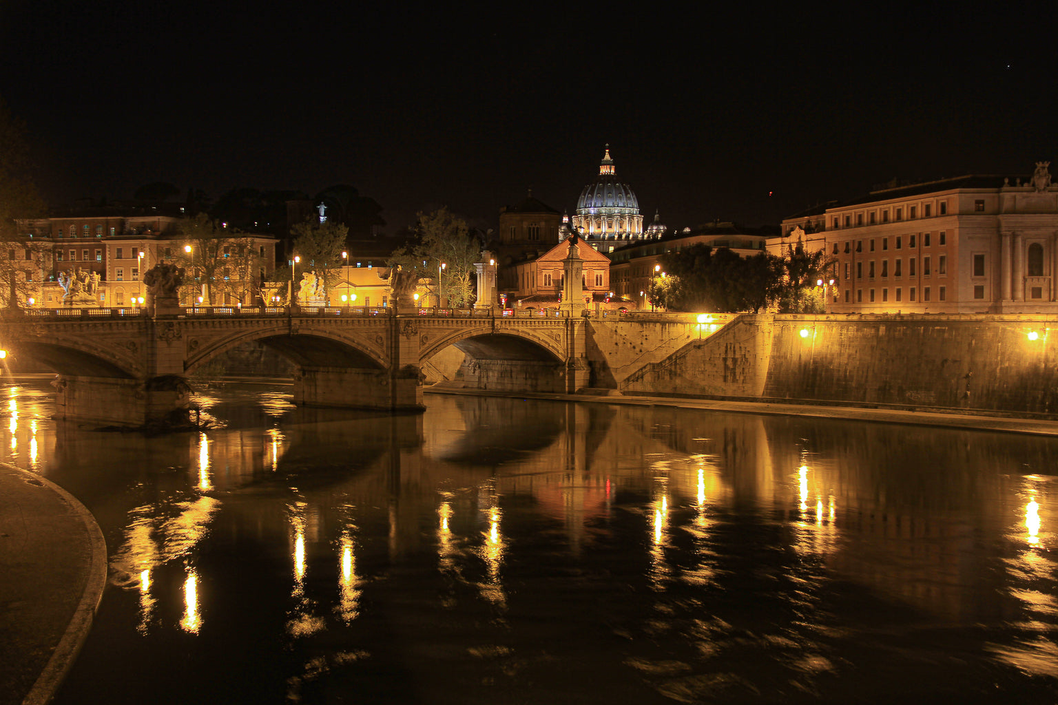 City bridge and warm lights reflected on a river at night with Vatican done at the background.
