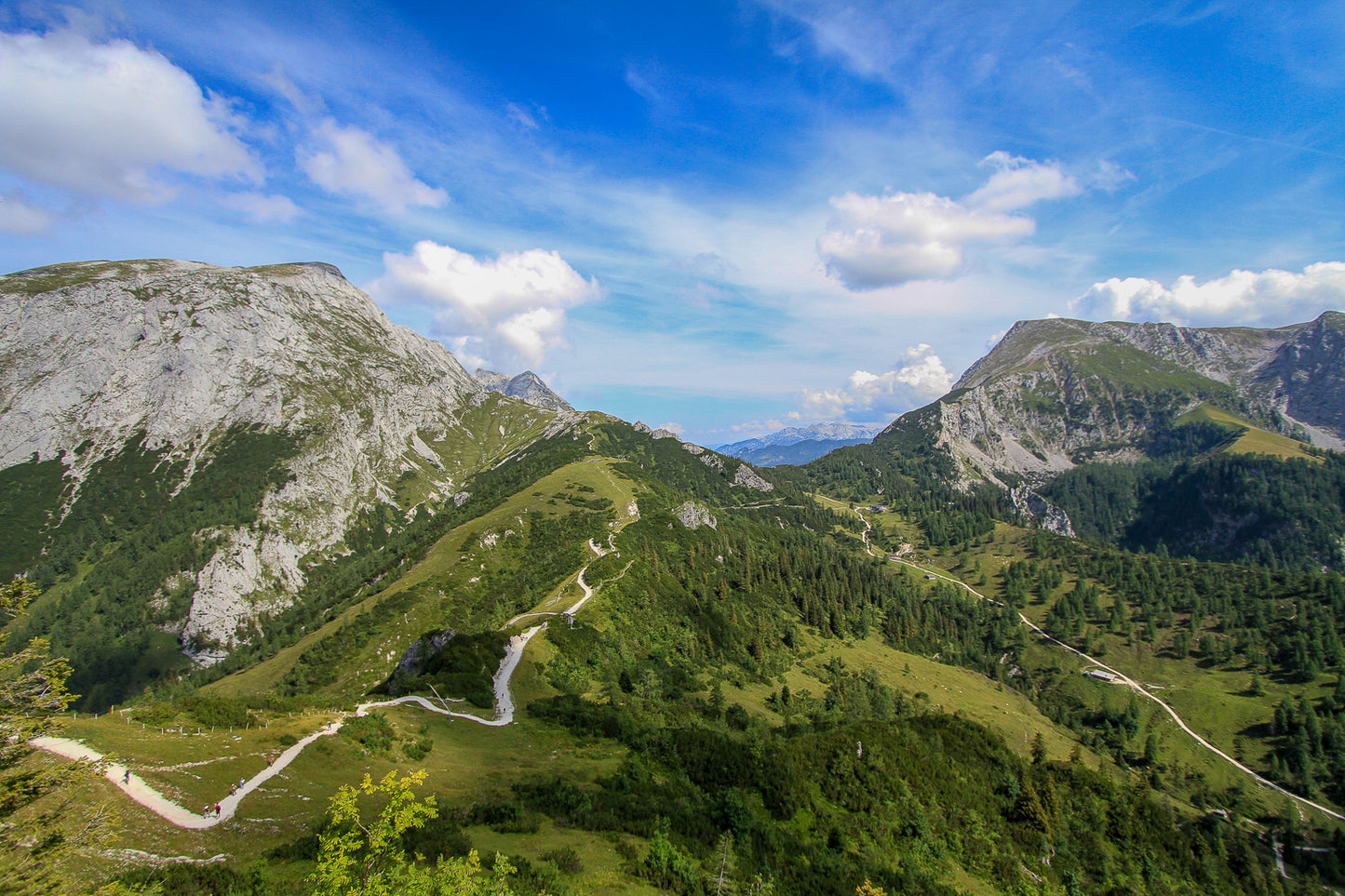 Mountain ridge landscape with winding trails, green slopes, and a bright blue sky with clouds.