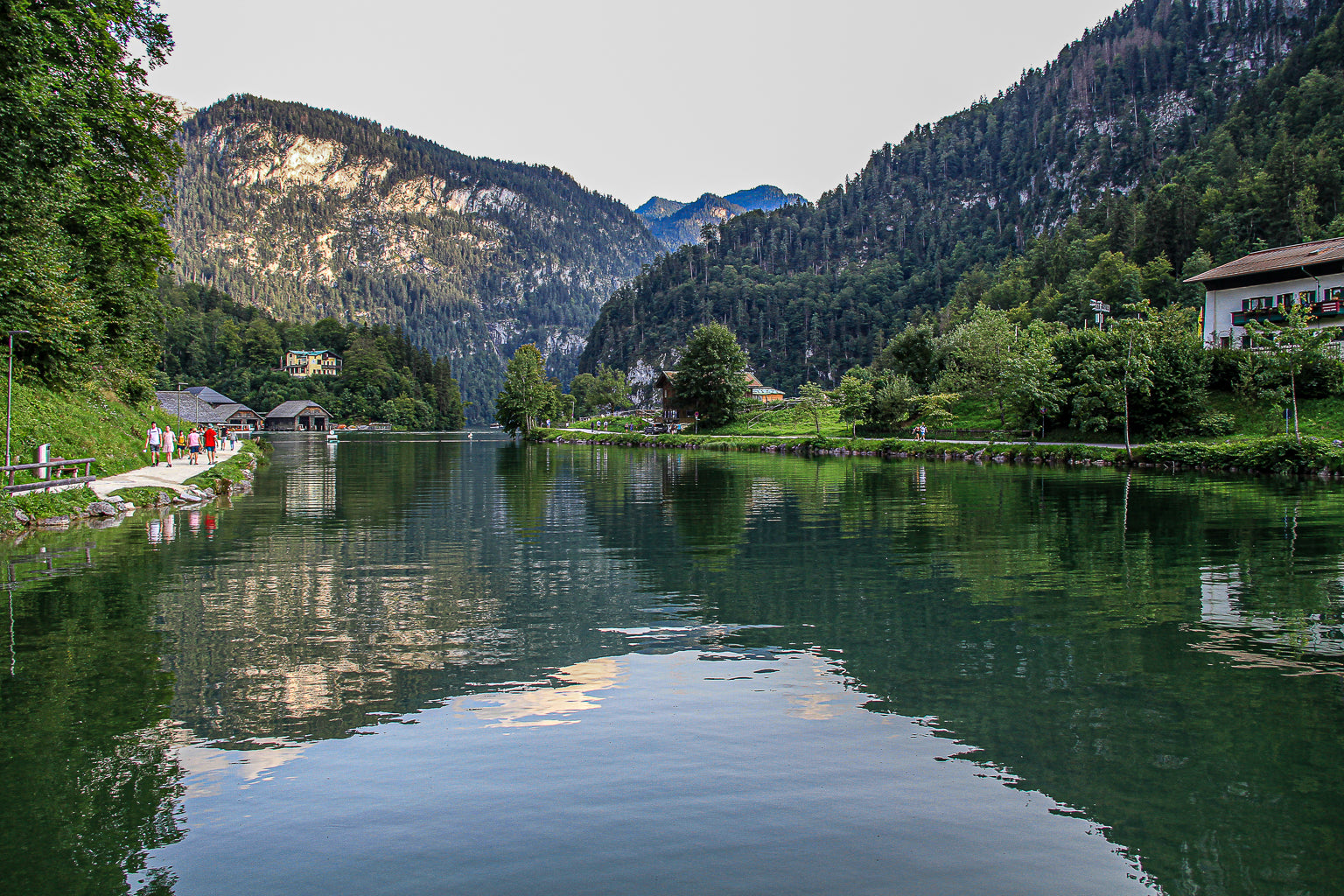 Reflective mountain waterway at Konigsee with green banks, distant peaks, and people walking along a lakeside path.