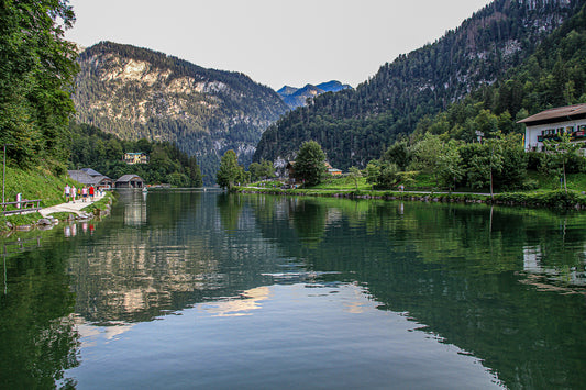 Reflective mountain waterway at Konigsee with green banks, distant peaks, and people walking along a lakeside path.