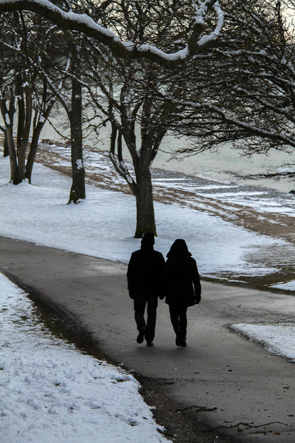 Two people walking on a snowy park path beneath bare trees.