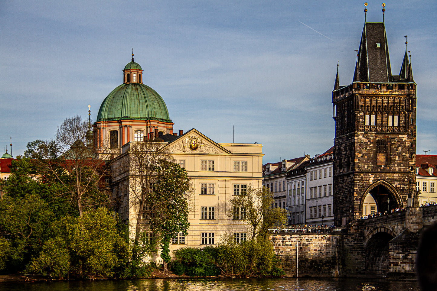 Historic stone bridge tower beside a river, with a green-domed church/building under a clear sky in a European city.