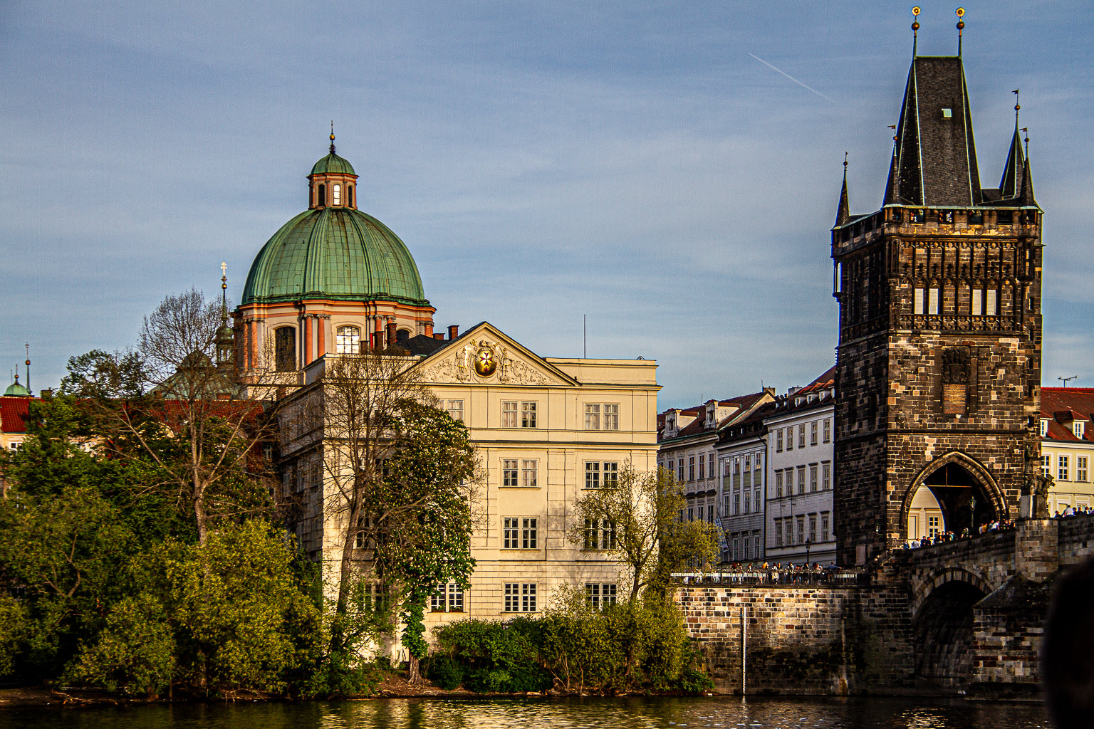 Historic stone bridge tower beside a river, with a green-domed church/building under a clear sky in a European city.