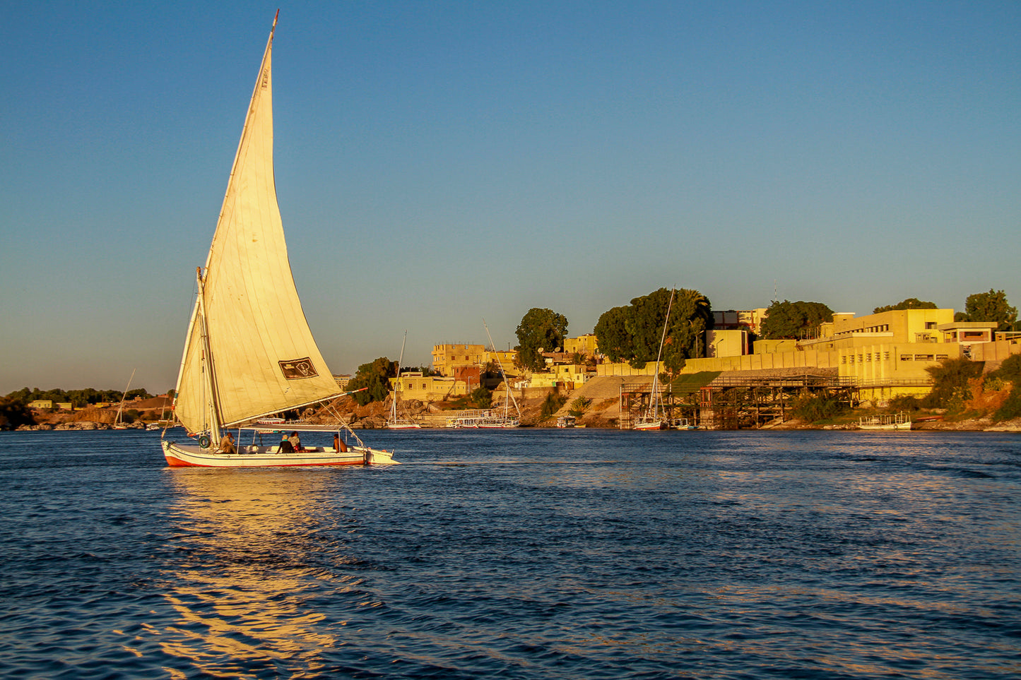 White-sailed boat cruising on a calm river at sunset with a shoreline town in the background.