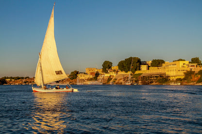 White-sailed boat cruising on a calm river at sunset with a shoreline town in the background.