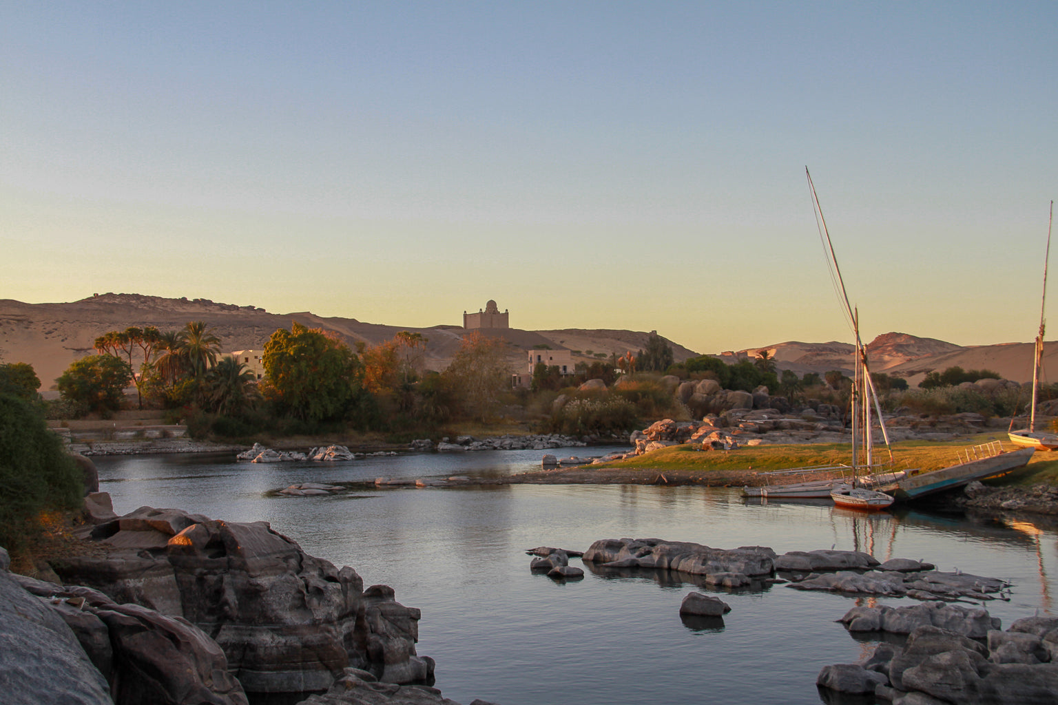 Quiet river bend at sunset with moored sailboats, rocks, palms, and distant desert hills.