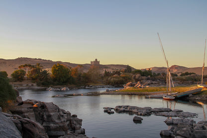 Quiet river bend at sunset with moored sailboats, rocks, palms, and distant desert hills.